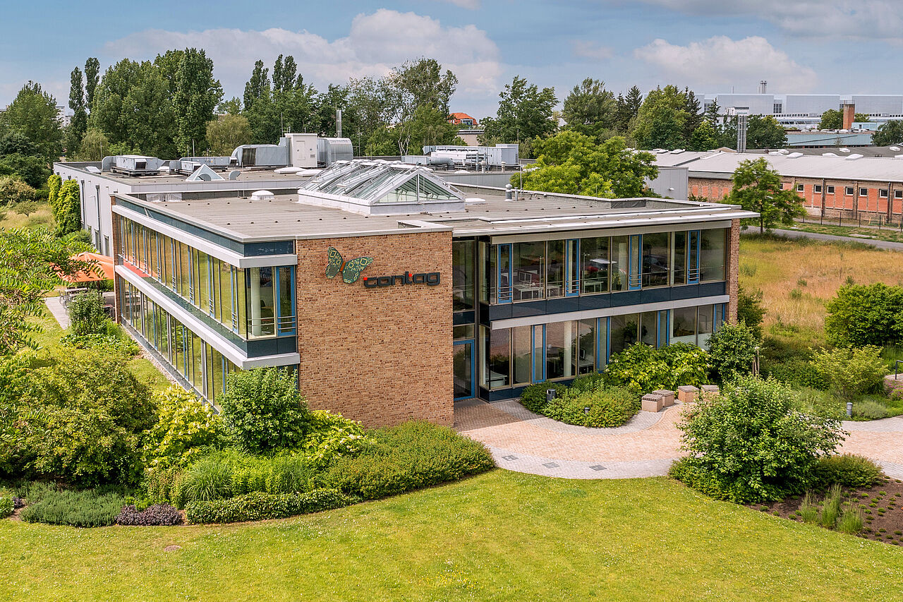 Außenansicht der CONTAG AG in Berlin: Das moderne Firmengebäude mit Klinkerfassade und Glasfront, eingebettet in eine gepflegte Grünanlage mit Gartenwegen.