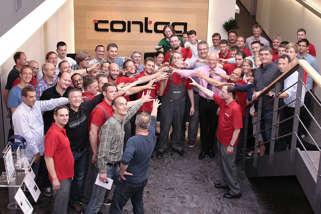 Gemeinsamer Erfolg bei CONTAG: Die Belegschaft versammelt sich im Foyer unter dem Firmenlogo und feiert eine Auszeichnung, wobei alle Teammitglieder symbolisch auf den Award in der Mitte zeigen.