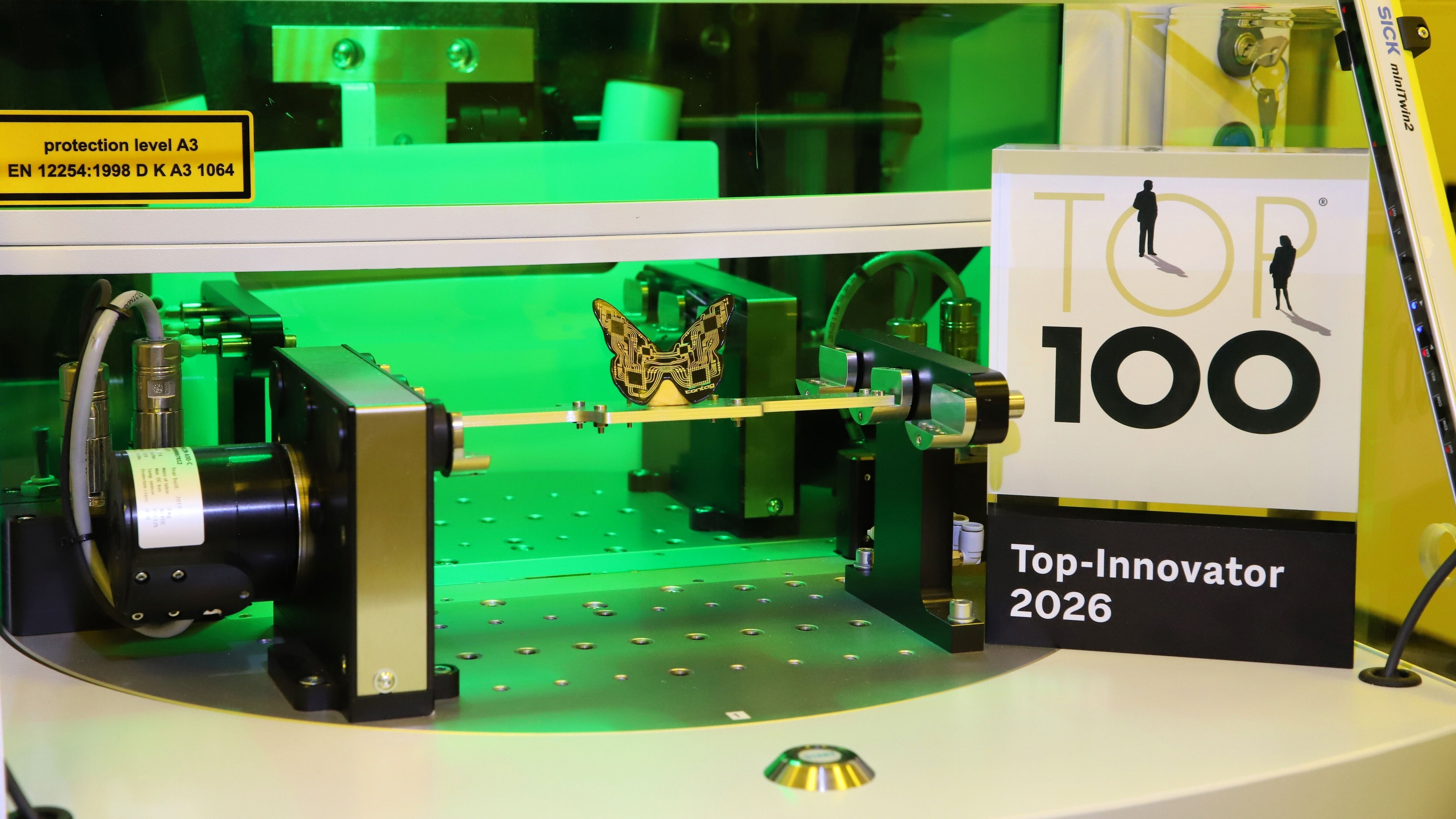Auszeichnung für CONTAG: Das Siegel „Top 100 Innovator 2026“ neben einer hochpräzisen Fertigungsanlage, in der ein filigraner 3D-MID Leiterplatten-Schmetterling bearbeitet wird.