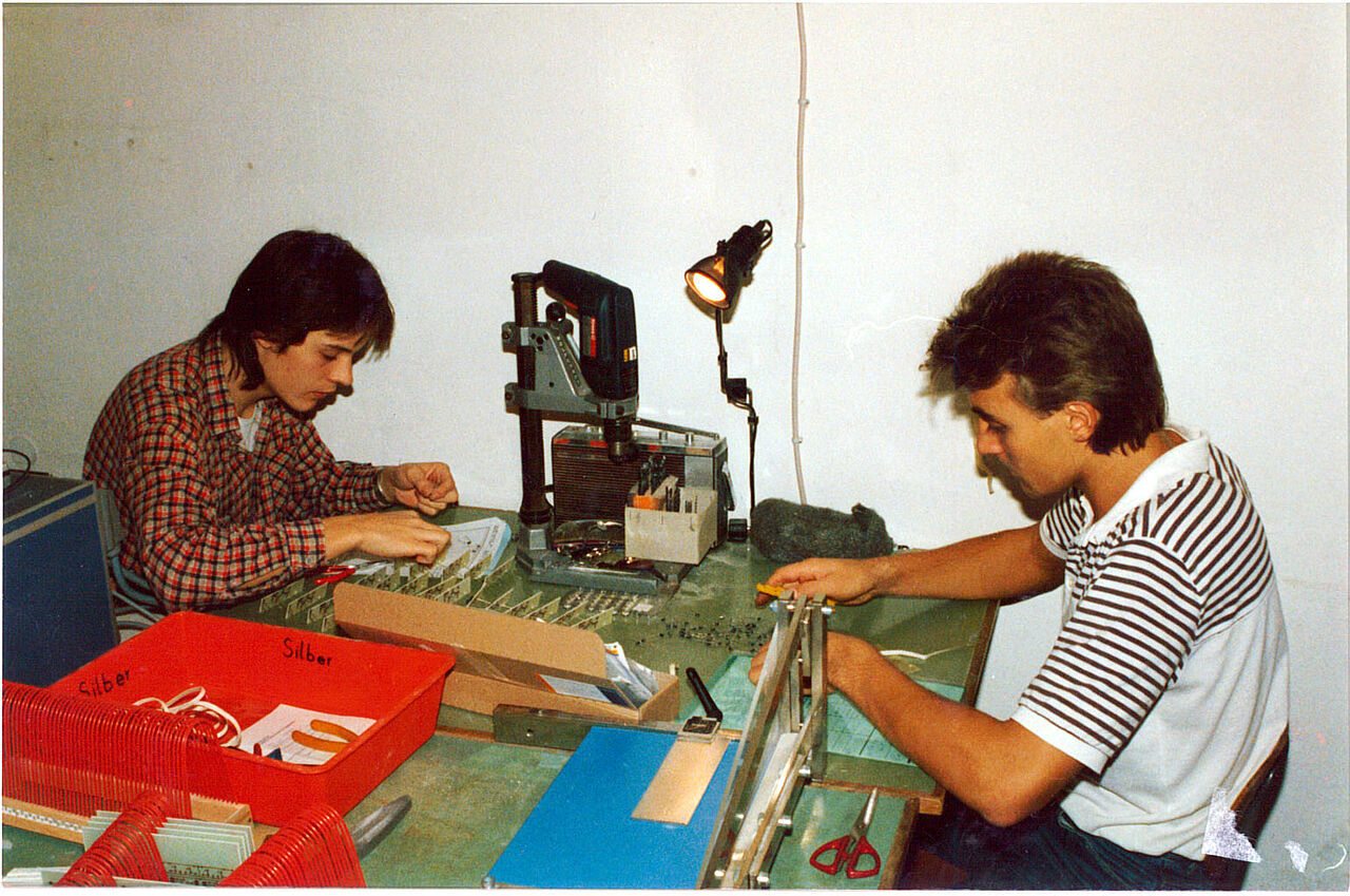 Blick in die frühen Jahre der CONTAG Leiterplatten-Fertigung: Zwei junge Männer arbeiten konzentriert an einem einfachen Werkzeugtisch mit Handbohrmaschine und manuellen Schneidewerkzeugen.