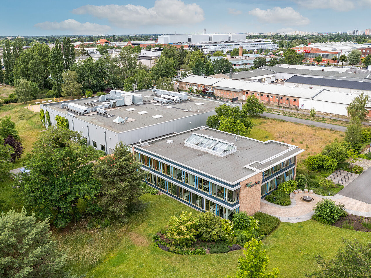 Vogelperspektive auf das Firmengelände der CONTAG AG in Berlin: Luftbild des modernen Verwaltungsgebäudes und der angrenzenden Produktionshallen, umgeben von weitläufigen Grünflächen.