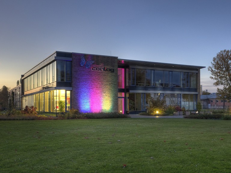 Abendstimmung bei der CONTAG AG in Berlin: Das moderne Firmengebäude mit beleuchteter Glasfront und farbiger Licht-Illumination in Blau und Pink vor einem weitläufigen Rasen.