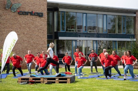 Aktives Betriebliches Gesundheitsmanagement bei der CONTAG AG: Eine Gruppe von Mitarbeitern in roten Firmen-Shirts macht gemeinsam mit einer Trainerin Fitness- und Dehnübungen auf dem Rasen vor dem Firmengebäude.
