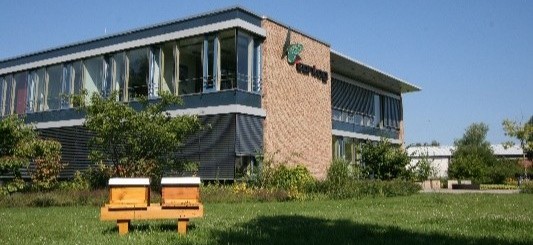 Gelebter Umweltschutz bei CONTAG: Zwei hölzerne Bienenstöcke stehen auf einer grünen Blumenwiese direkt vor dem modernen Hauptgebäude der Firmenzentrale in Berlin.