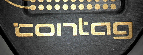 Nahaufnahme des goldenen CONTAG-Logos, das mittels 3D-MID-Technologie auf ein schwarzes Duroplast-Material aufgebracht wurde, um die Eignung spezieller Materialien zu demonstrieren.