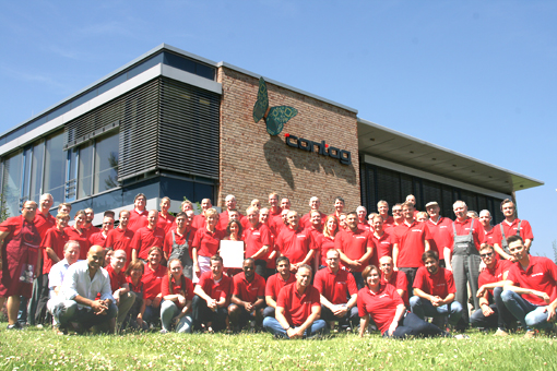 Das Team der CONTAG AG in Berlin: Gruppenfoto der Belegschaft in roten Firmentrikots vor dem modernen Unternehmensgebäude unter blauem Himmel.
